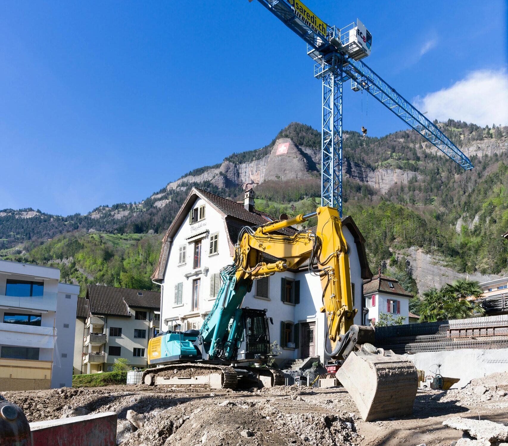 Scenic construction site with a crane and excavator in a Swiss alpine village under clear blue skies.